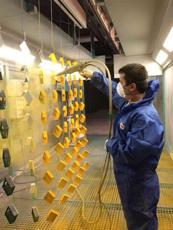 Produktteile werden gelb beschichtet Ein Mitarbeiter beschichtet mit einem Handsprühgerät die aufgehängten Produktteile mit gelber Farbe.